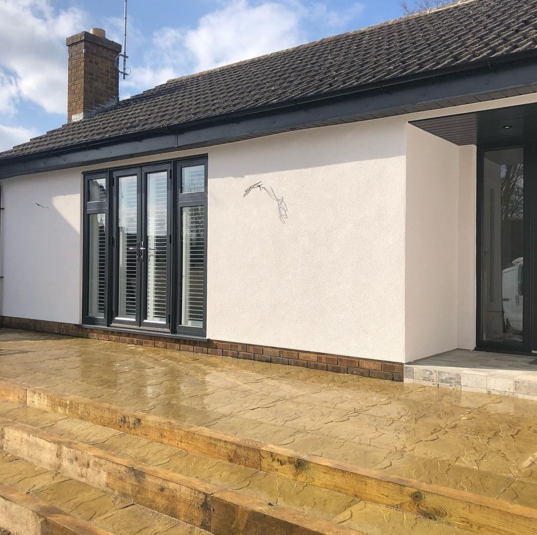 Single-storey extension finished in off-white render with grey glazed doors and a new patio